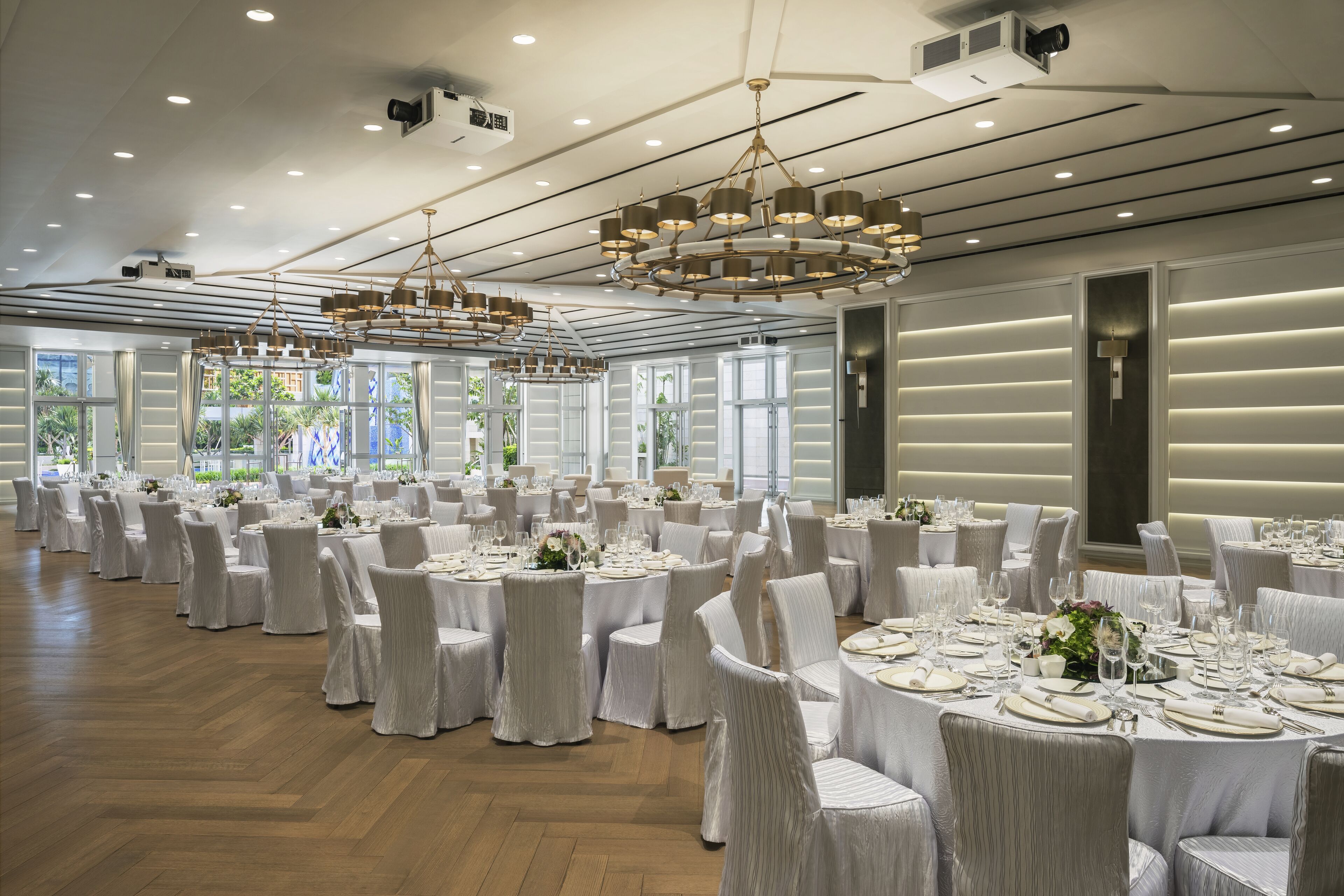 Une grande salle de banquet avec des tables rondes dressées pour le repas, des chaises blanches et un lustre moderne.