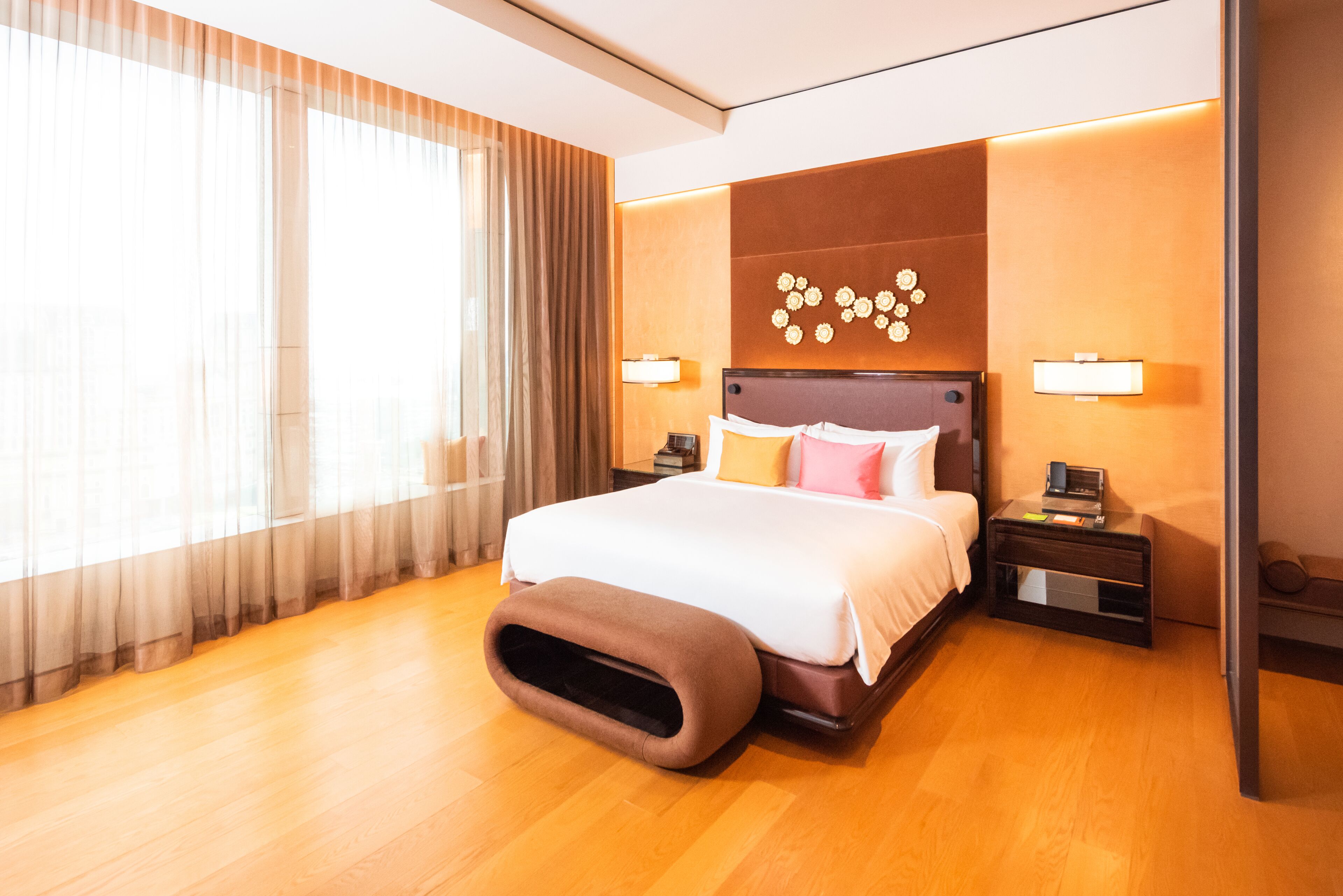A hotel room with a large bed, two bedside tables, a nightstand, a wall-mounted lamp, and a window with sheer curtains.