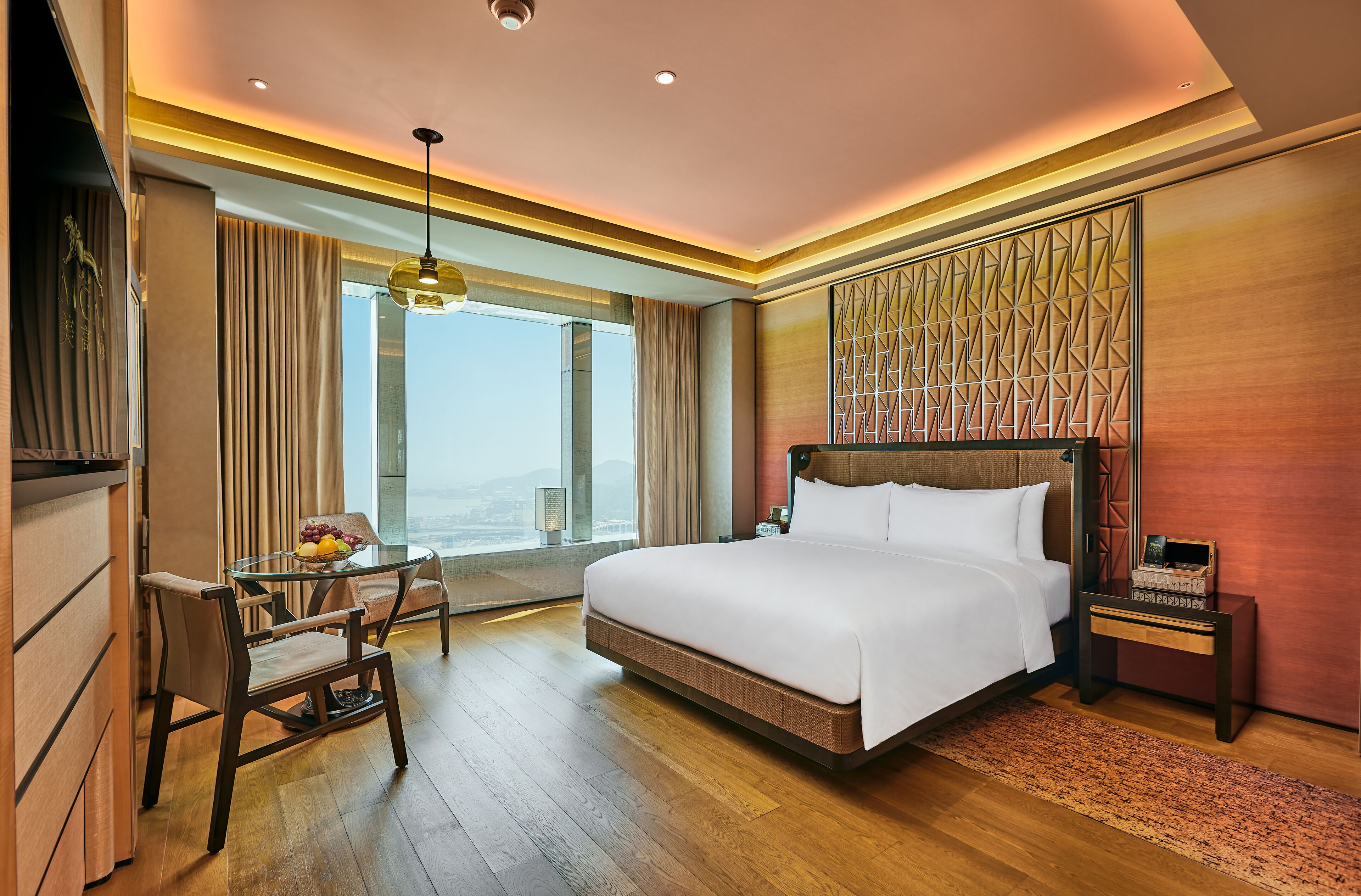 A hotel room with a large bed, a desk with a chair, a small table with fruit, and a view of the city.