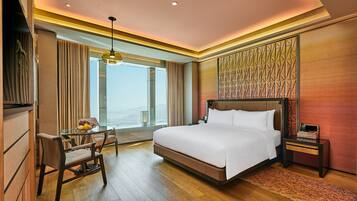 A hotel room with a large bed, a desk with a chair, a small table with fruit, and a view of the city.