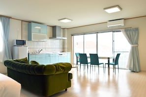 Een moderne woonkamer met een groene bank, een eethoek met blauwgroene stoelen, een keuken met blauwe kastjes en een groot raam met gordijnen.