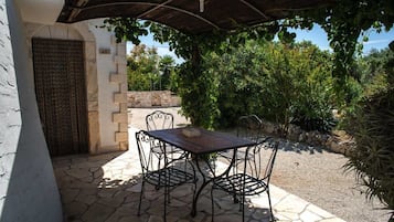 Un patio al aire libre con una mesa de piedra y sillas de metal, rodeado de follaje y un muro de piedra.