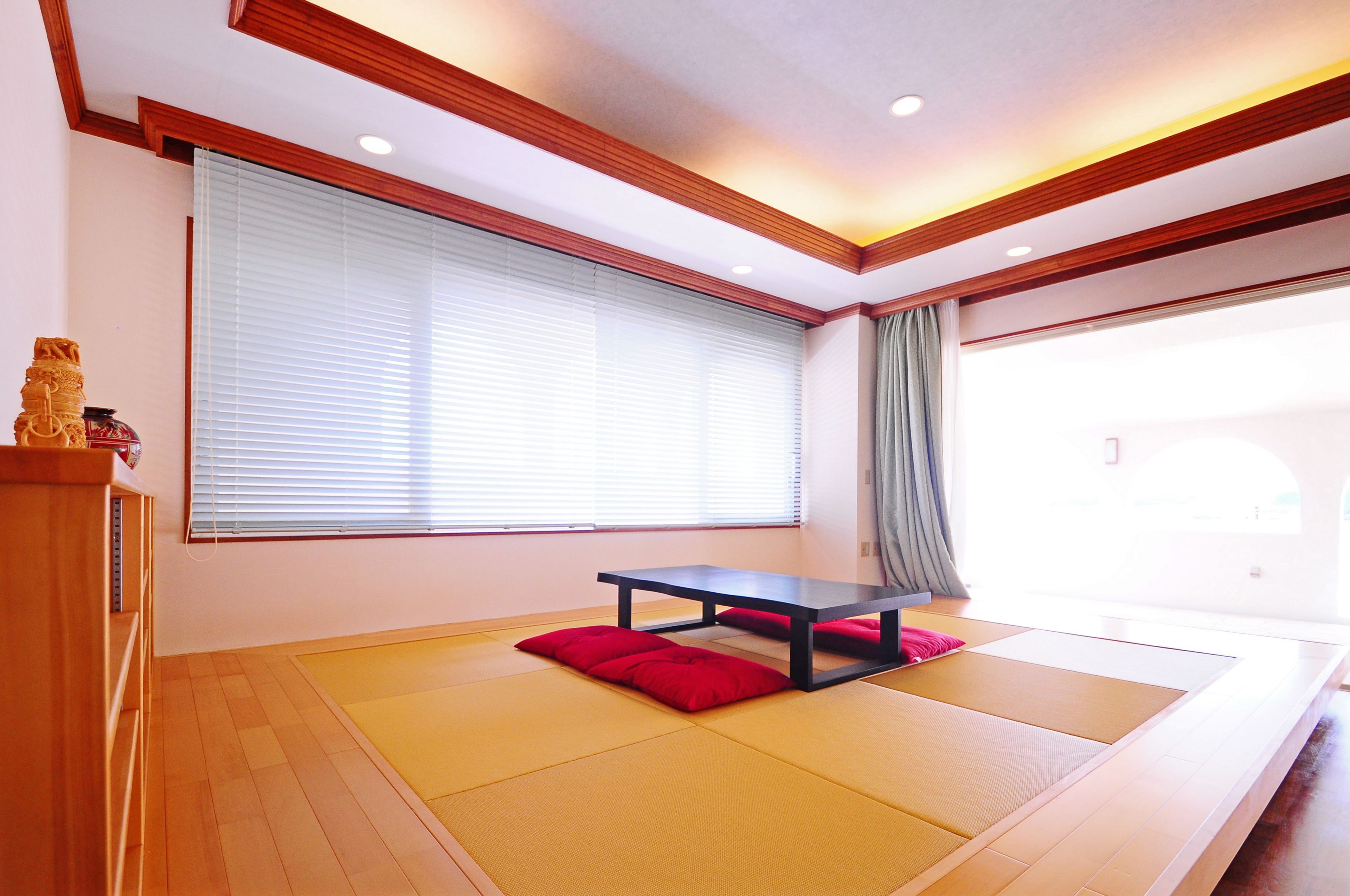A tatami mat room with a low table and cushions, wooden beams on the ceiling, and a large window with blinds.