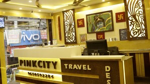 A travel desk with a reception area, including a counter with a phone and a computer, framed pictures on the wall, and a ceiling fan. - Hotel Pink City (New Delhi)