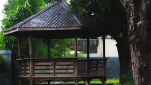 A wooden gazebo with a sloped roof, surrounded by lush greenery and a tree trunk.