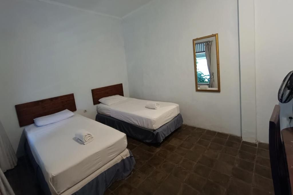A room with two single beds, a small mirror on the wall, and a fan on the right.