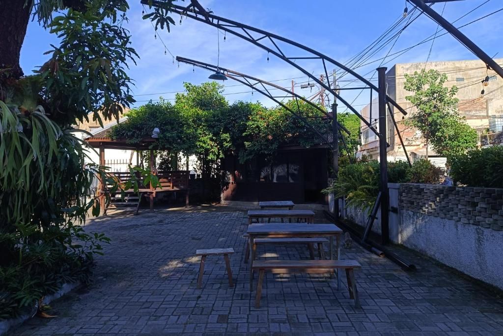 An outdoor patio with wooden tables and benches, surrounded by greenery and a pergola.