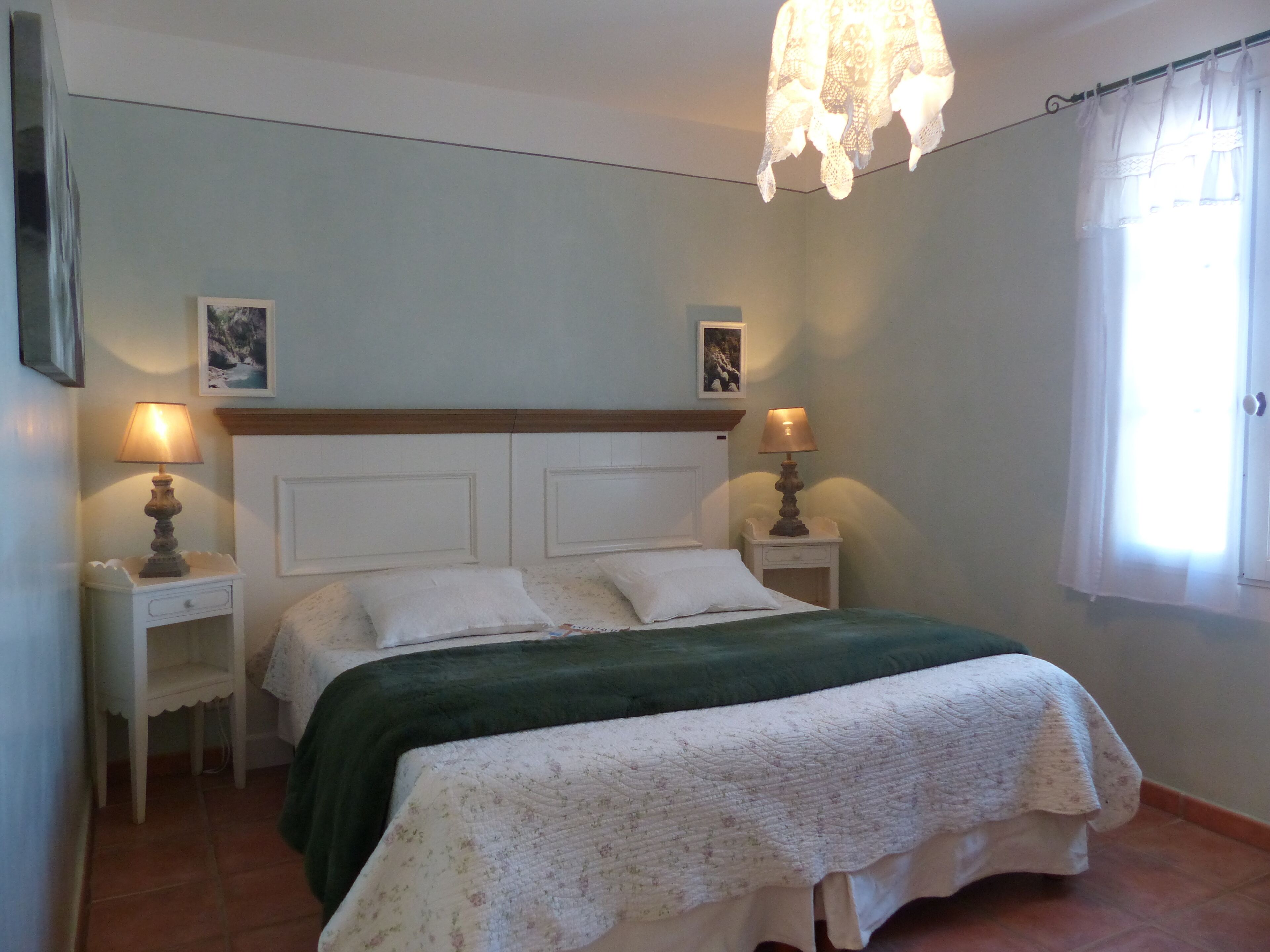 A bedroom with a bed, two bedside tables, a hanging light fixture, and a window with curtains.