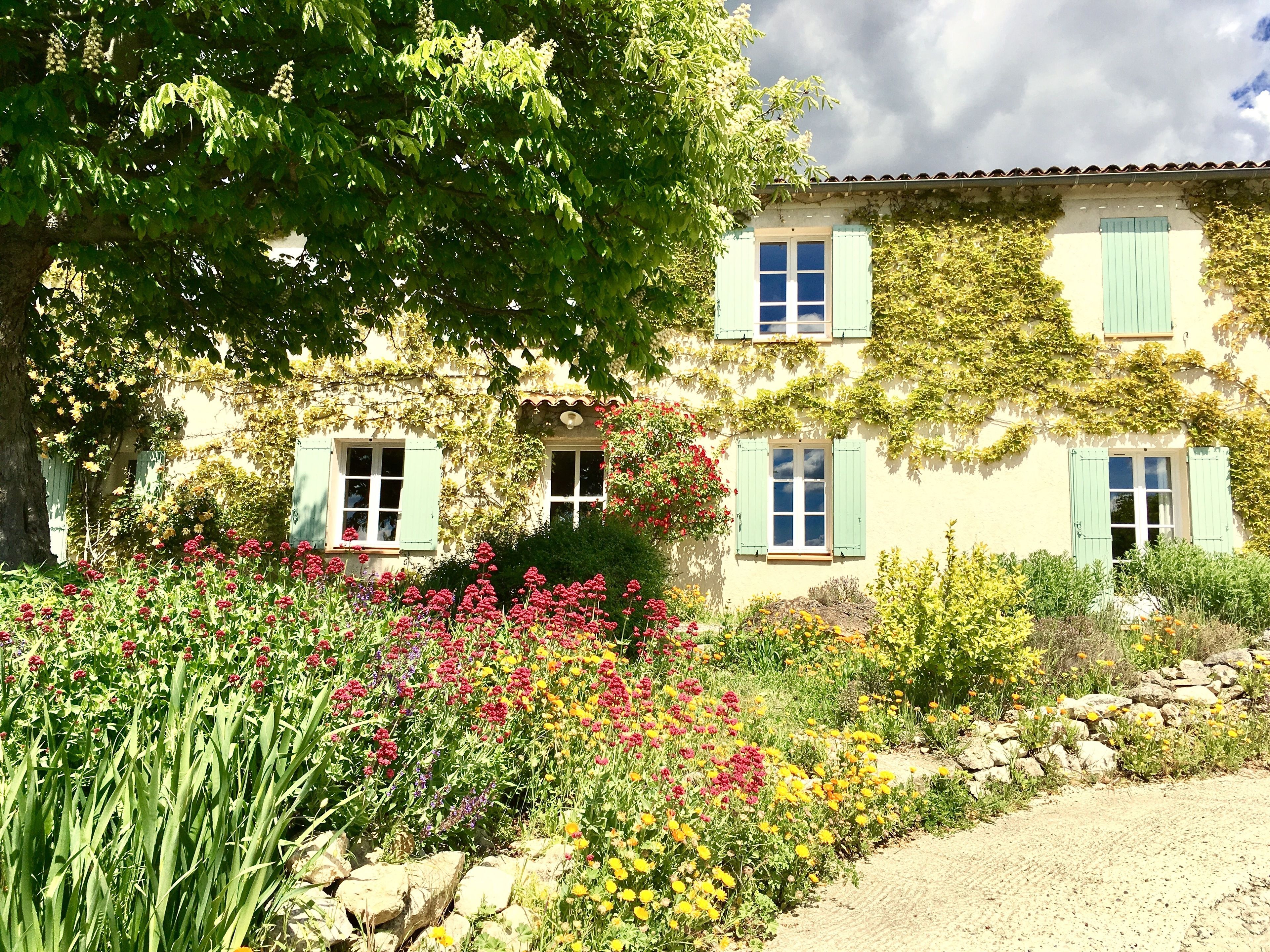 Un edificio de dos pisos con contraventanas verdes, rodeado de un jardín con diversas flores y un árbol grande.