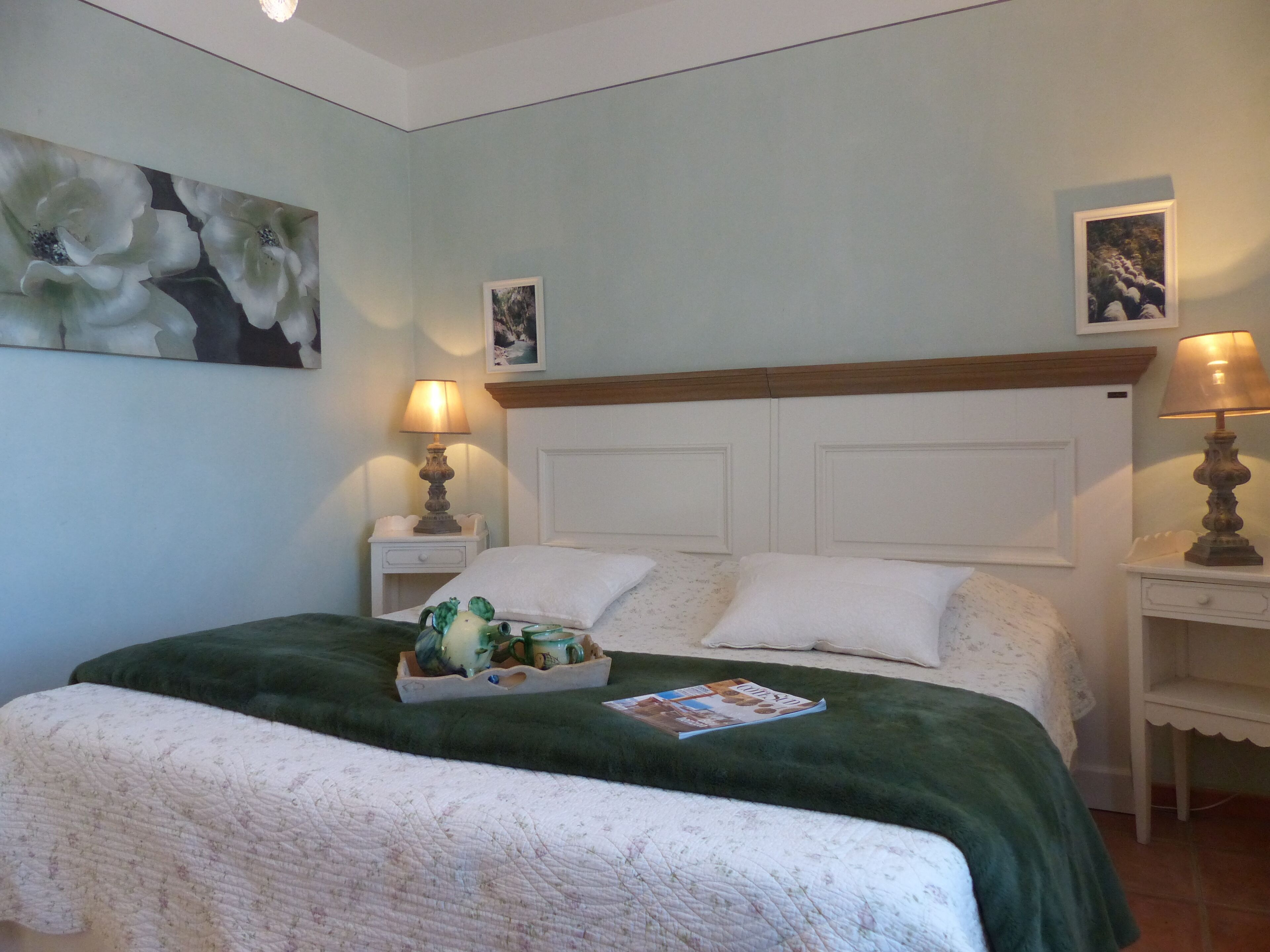 A neatly made bed with a green blanket, two bedside tables with lamps, and a wall with floral artwork.