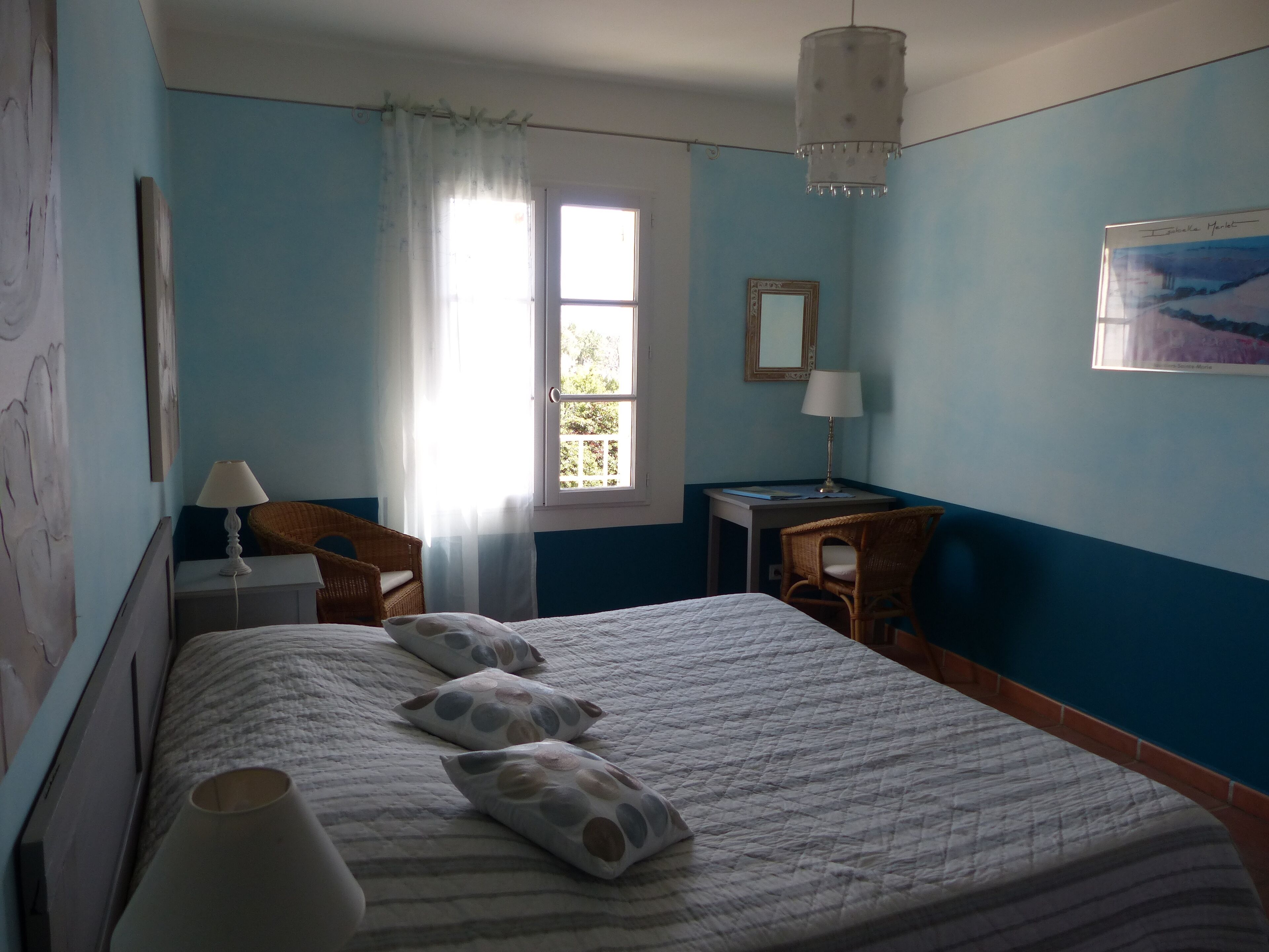 A bedroom with a bed, two chairs, a desk, a window with curtains, a hanging lamp, and a framed picture on the wall.