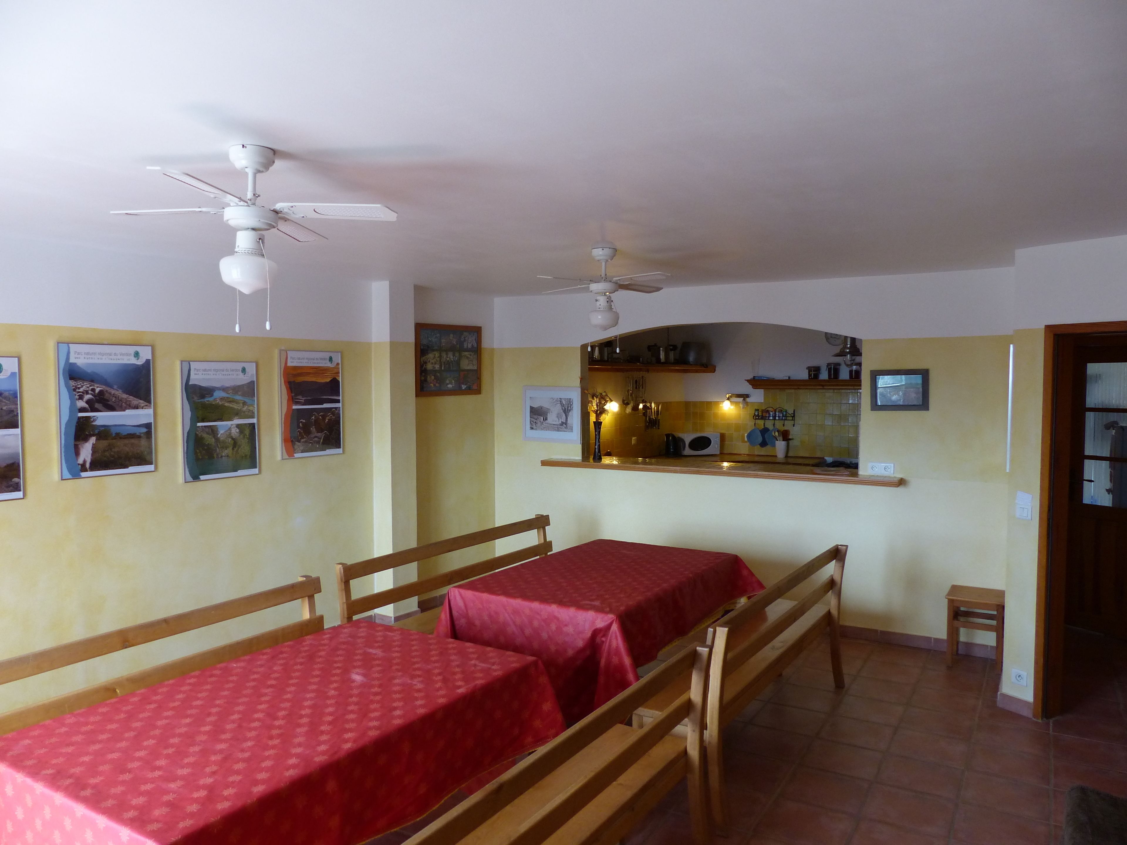 Un espace repas avec deux tables recouvertes de nappes rouges, des bancs en bois, un ventilateur de plafond et un coin cuisine visible à travers une arche.
