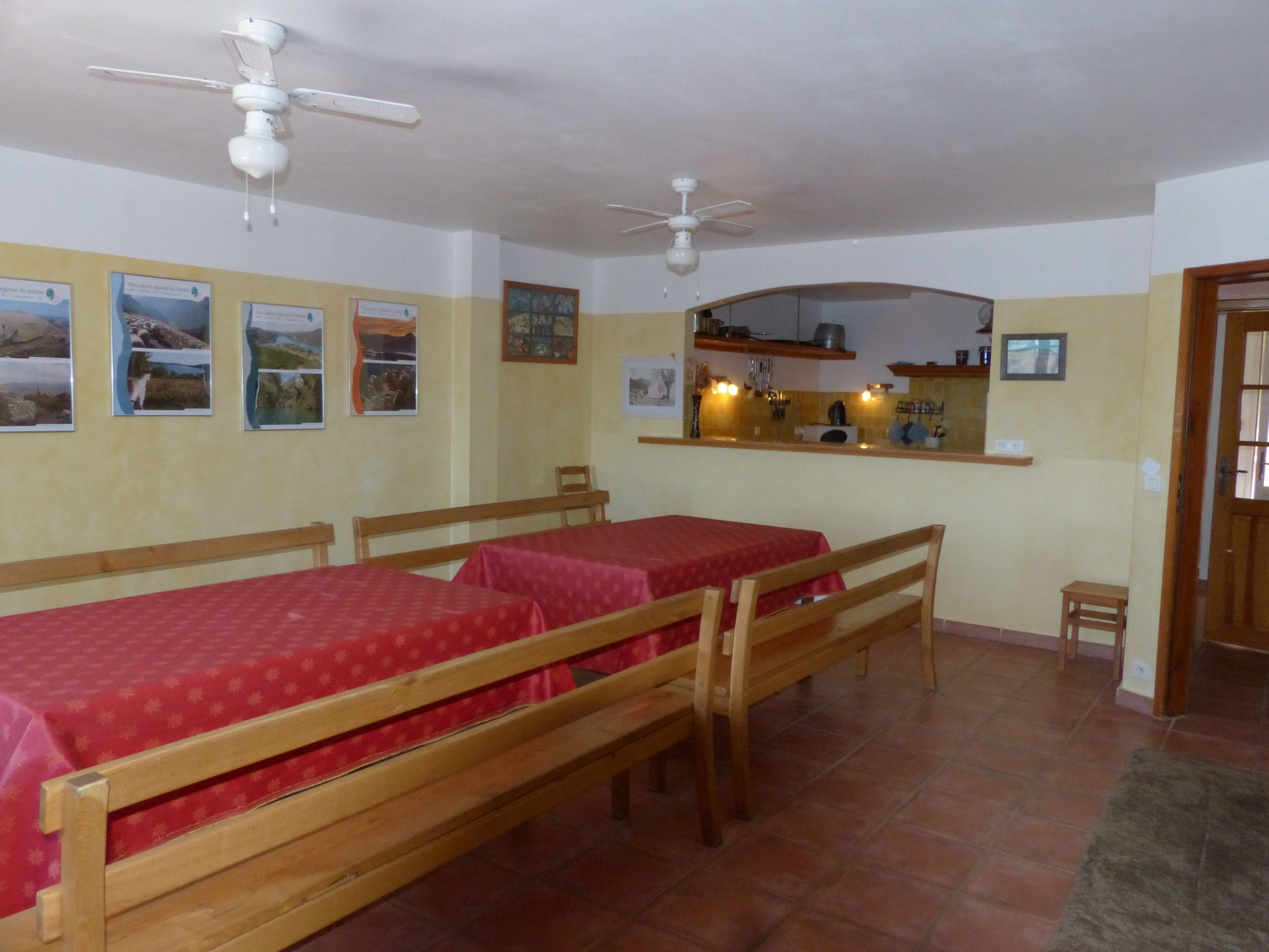 A room with two wooden beds with red bedding, a small kitchen area with a mirror above the counter, and a ceiling fan.