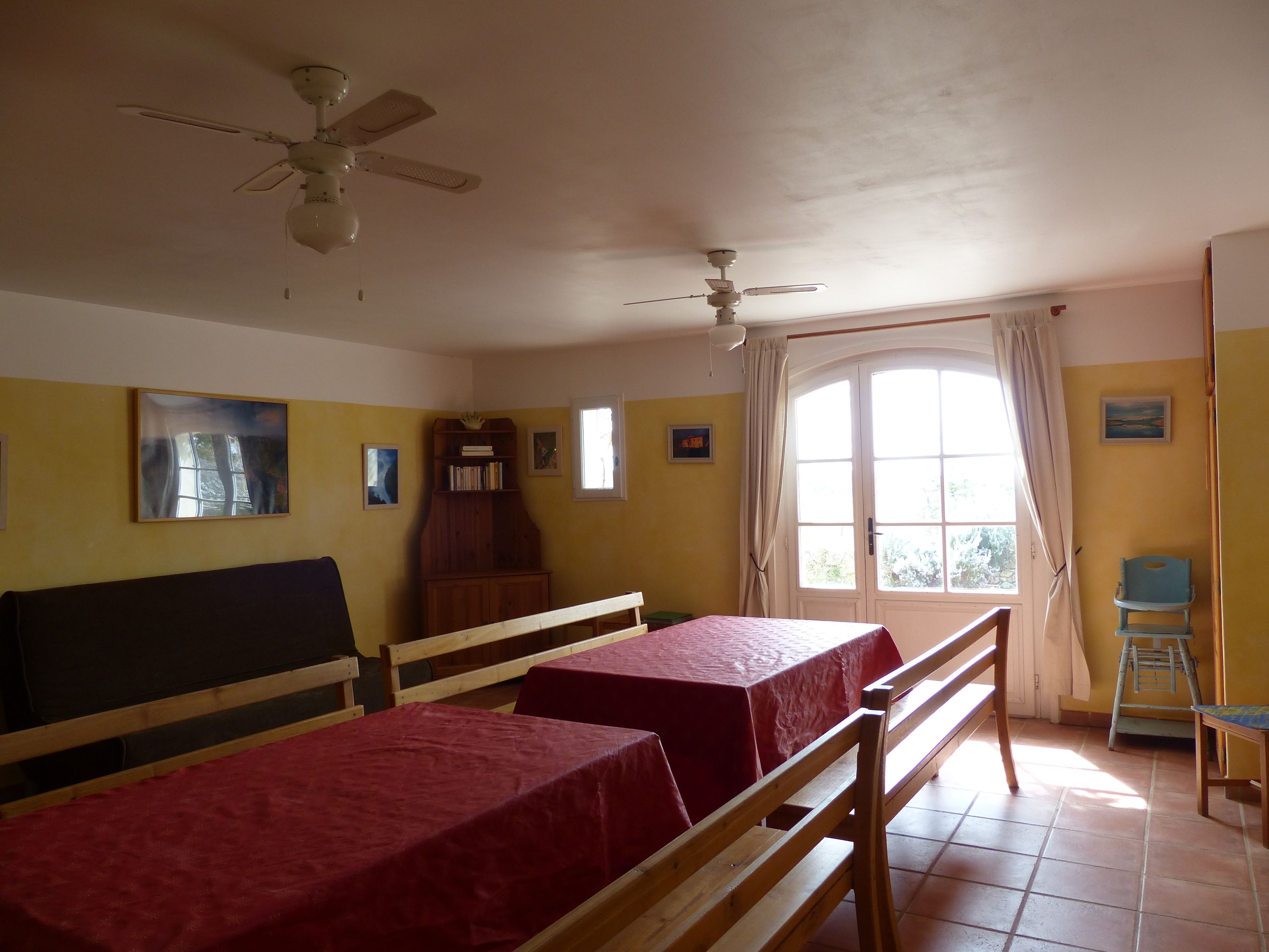 A room with two beds, a ceiling fan, a window with curtains, and a bookshelf.