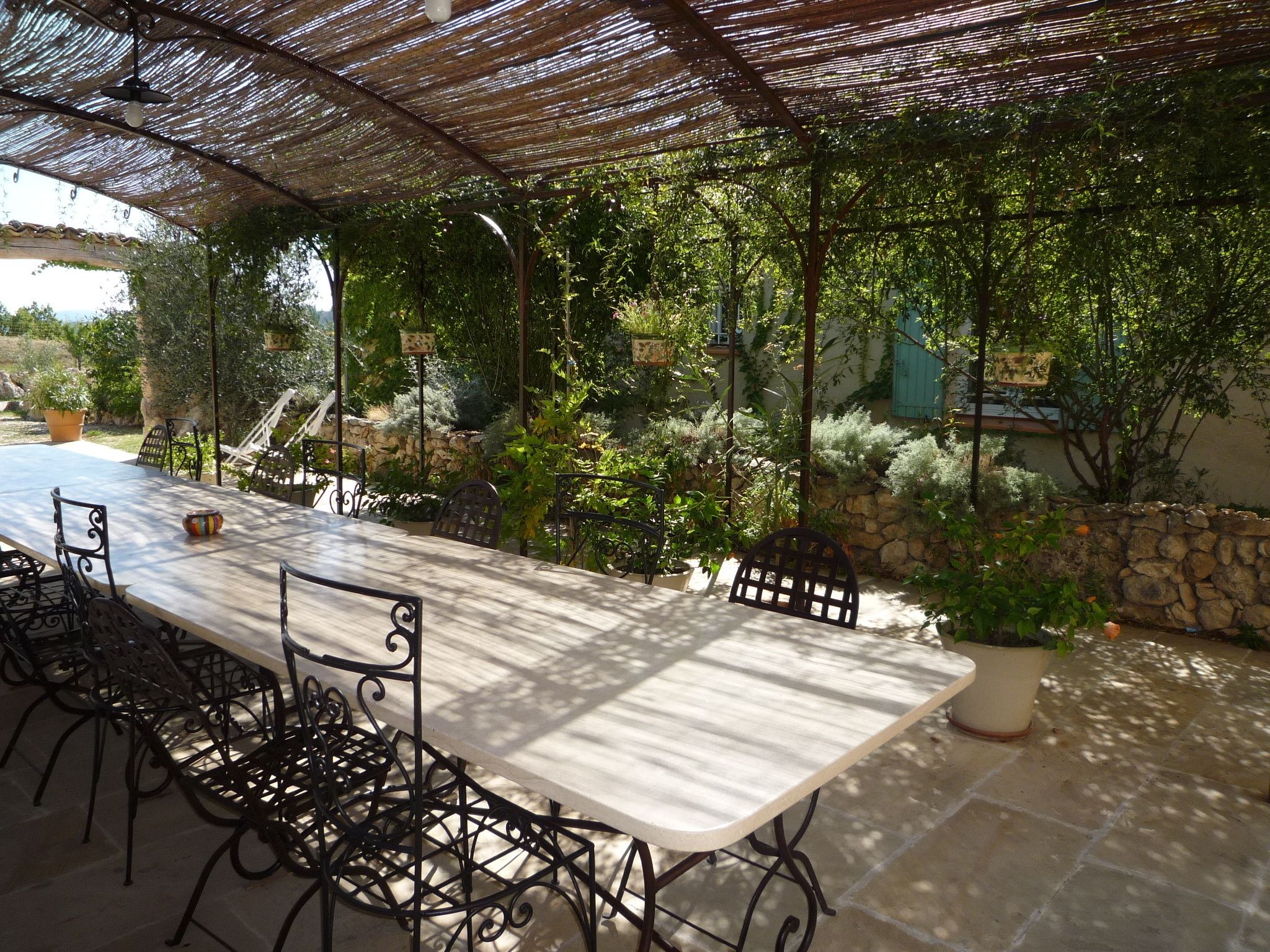 Un patio exterior con una mesa larga y sillas, rodeado de follaje y muros de piedra.