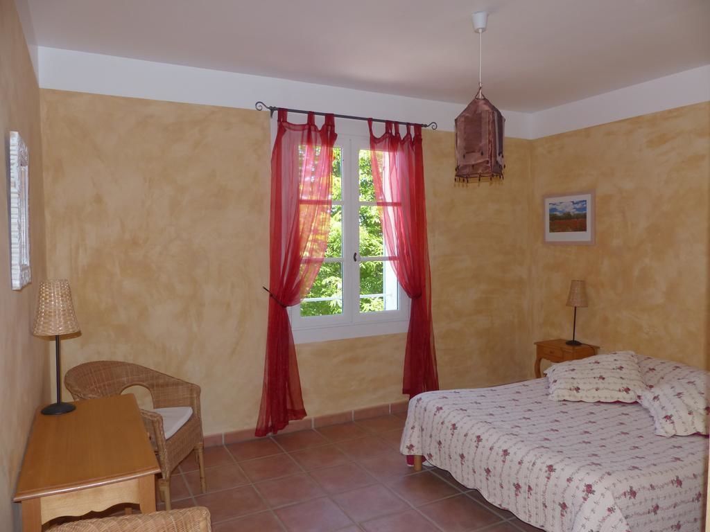 A bedroom with a bed, a chair, a table, a window with curtains, and a hanging light fixture.
