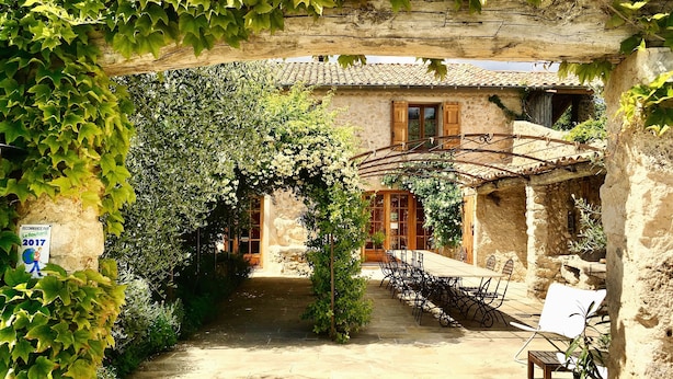 A rustic stone building with a tiled roof, a covered patio area with outdoor seating, and a garden path leading to the entrance. - La Ferme du Petit Ségriès (Moustiers-Sainte-Marie)