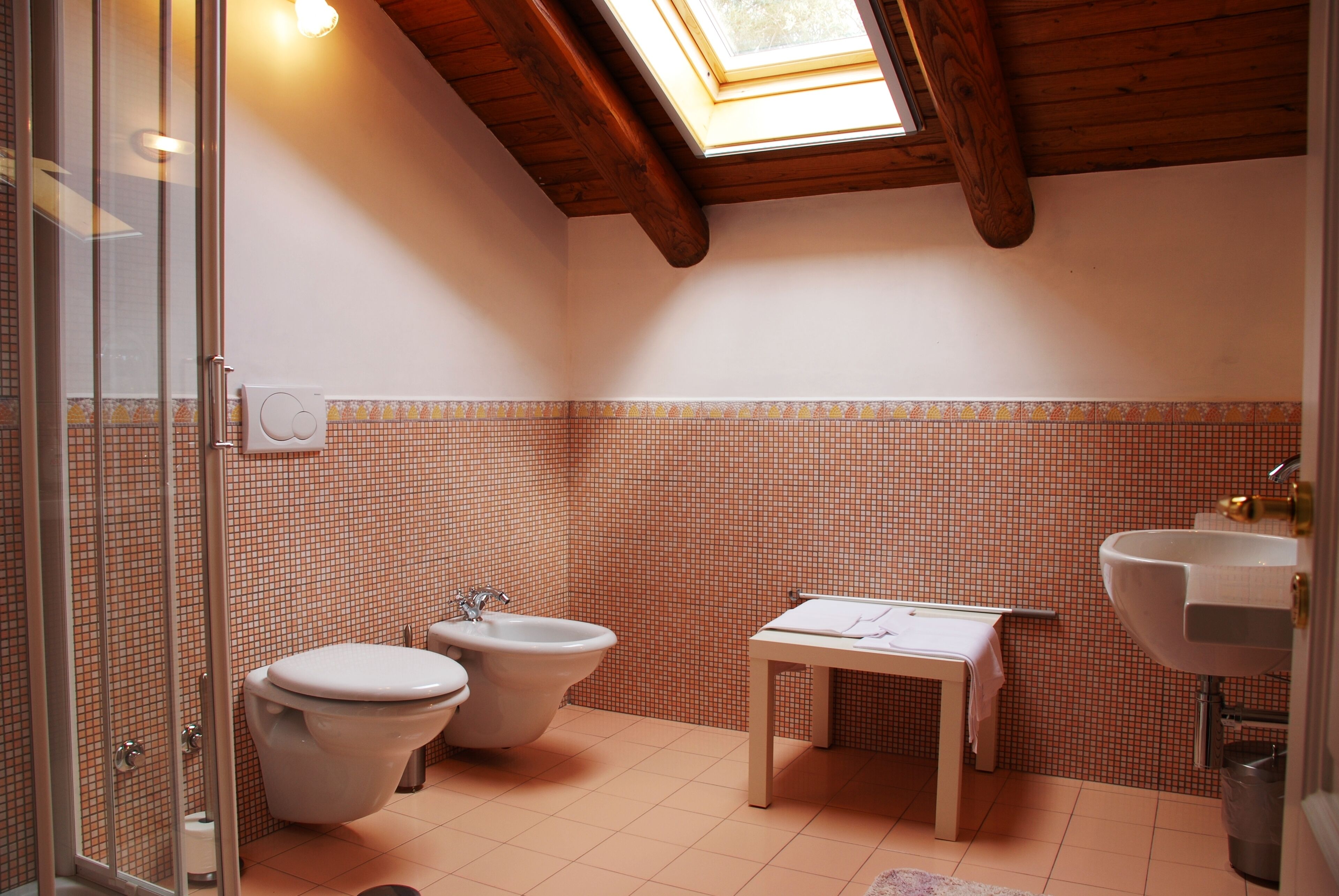 A bathroom with a skylight, two toilets, a bidet, a sink, and a small table with a towel.