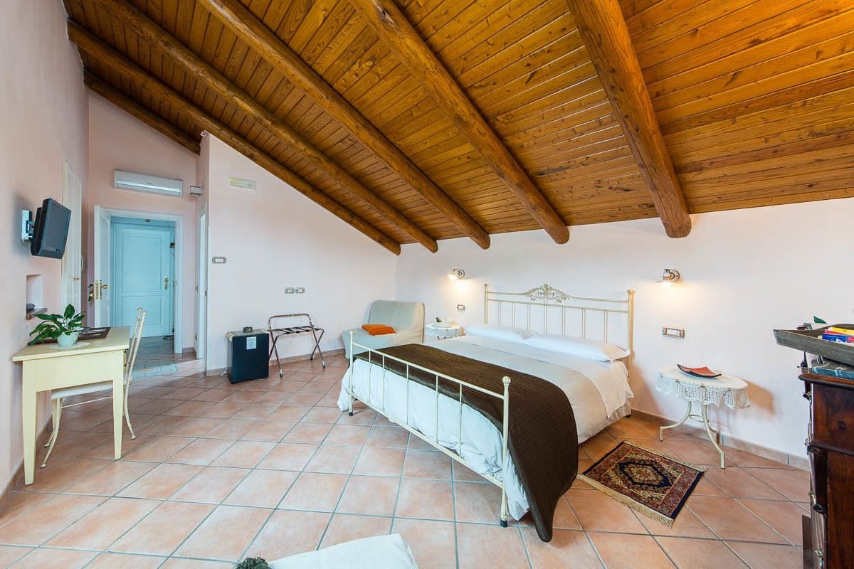Een slaapkamer met een houten plafond, een bed, een nachtkastje, een stoel, een klein tafeltje en een televisie aan de muur.