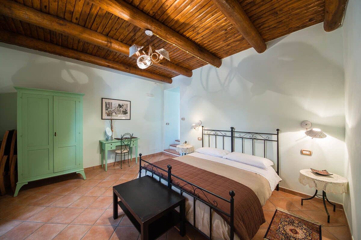 Een slaapkamer met een houten plafond, een bed met een bruine sprei, een groene kast, een bureau met stoel en een kleine tafel met lamp.