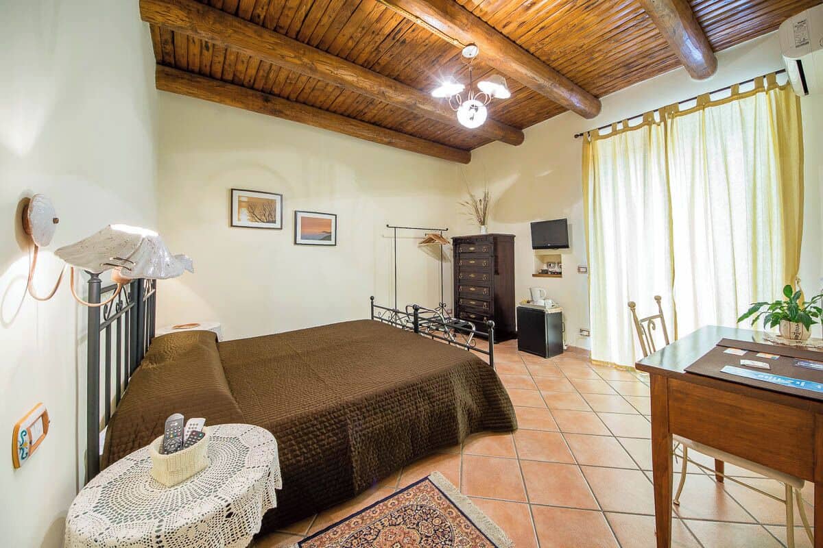 Een gezellige slaapkamer met een houten plafond, een bed met een bruine deken, een kleine tafel met een lamp en een bureau met een plantje.
