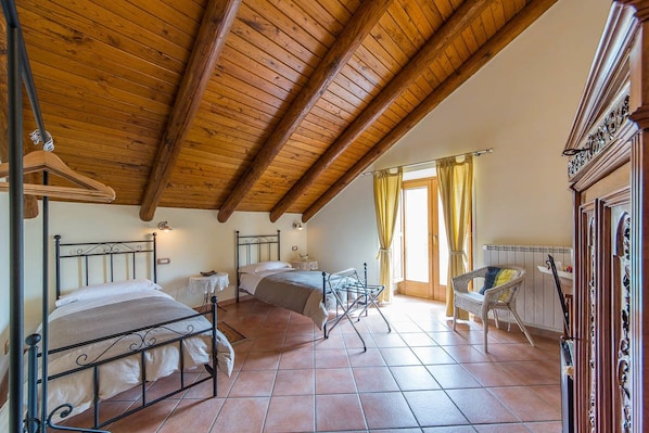 A bedroom with two beds, wooden ceiling beams, tiled floor, and a chair by the window.