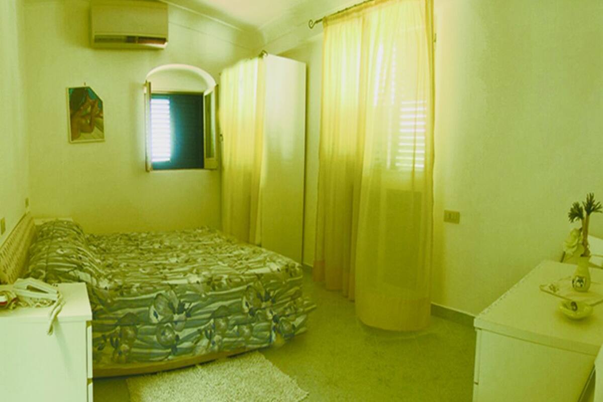 Un dormitorio con cama, mesita de noche, cómoda, ventana con cortinas y equipo de aire acondicionado.