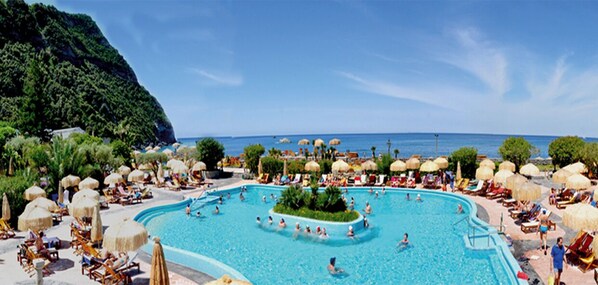 A large outdoor swimming pool surrounded by lounge chairs and umbrellas, with a view of the sea and a mountainous backdrop.