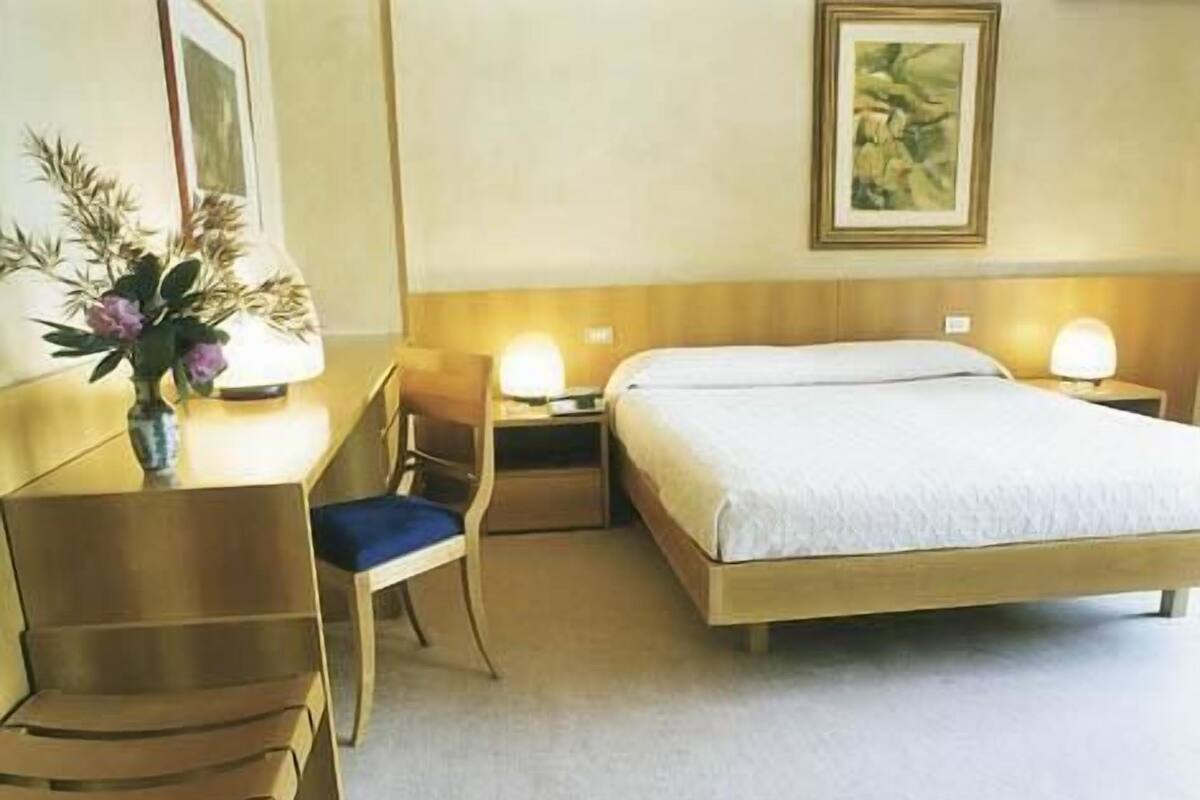 A hotel room with a bed, a desk, a chair, a nightstand, and a lamp.