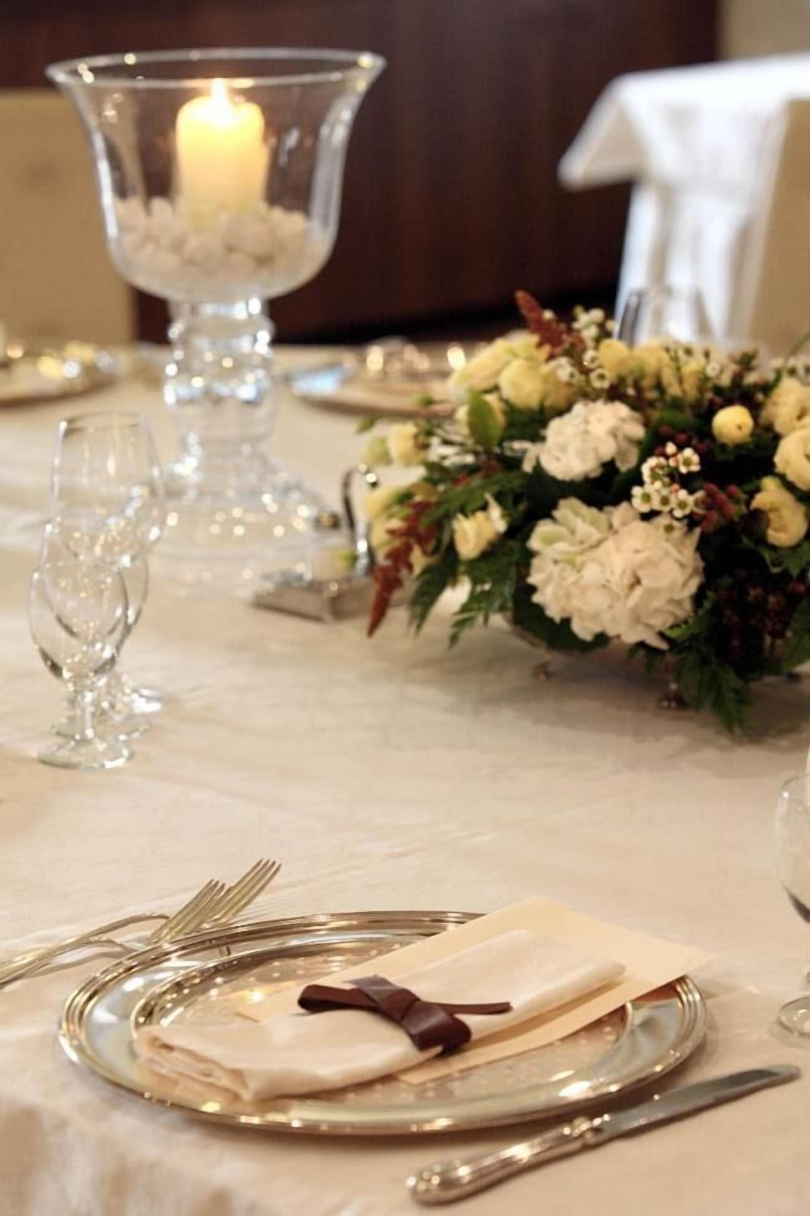 Una mesa preparada para cenar con una vela en un candelabro, un centro de mesa floral y un mantel doblado con una cinta marrón.