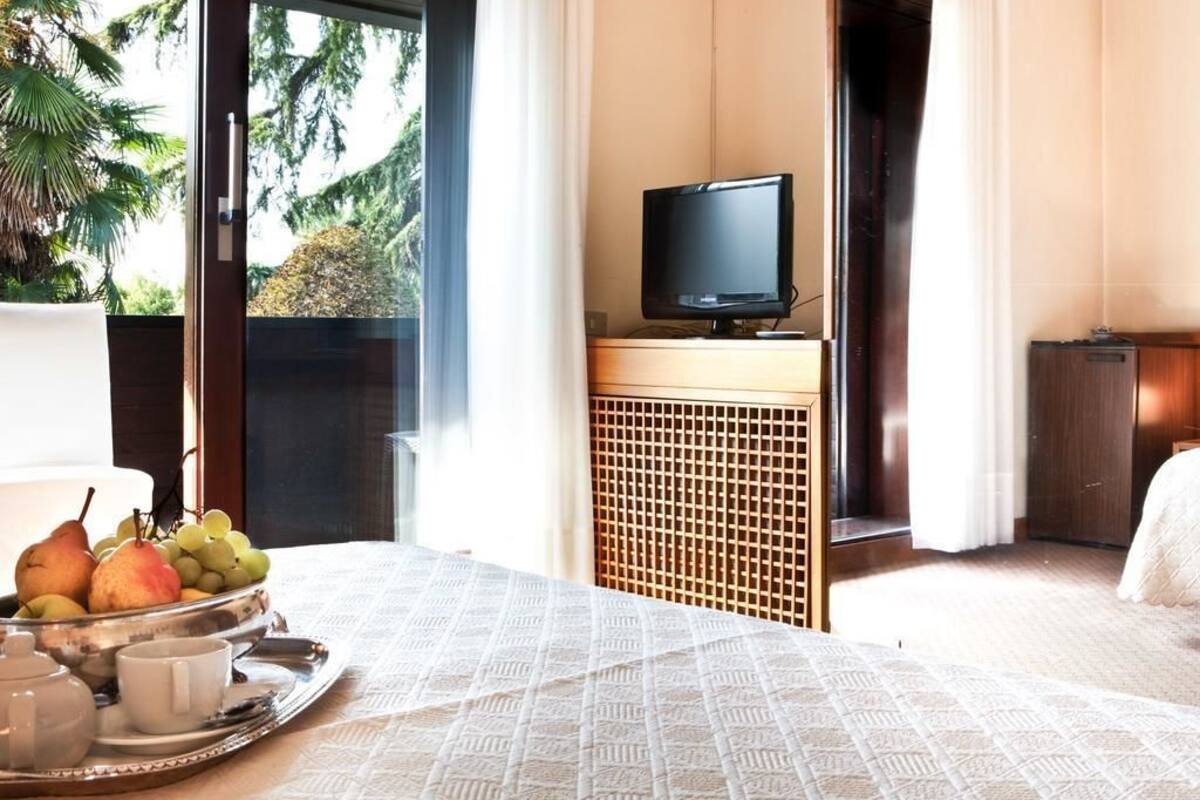 A hotel room with a bed, a television on a stand, a tray with fruits and a teapot, and a view of trees through the window.