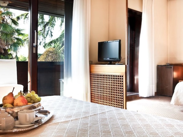 Una habitación de hotel con una cama, un televisor sobre un mueble, una bandeja con frutas y un hervidor, y vistas a árboles por la ventana.