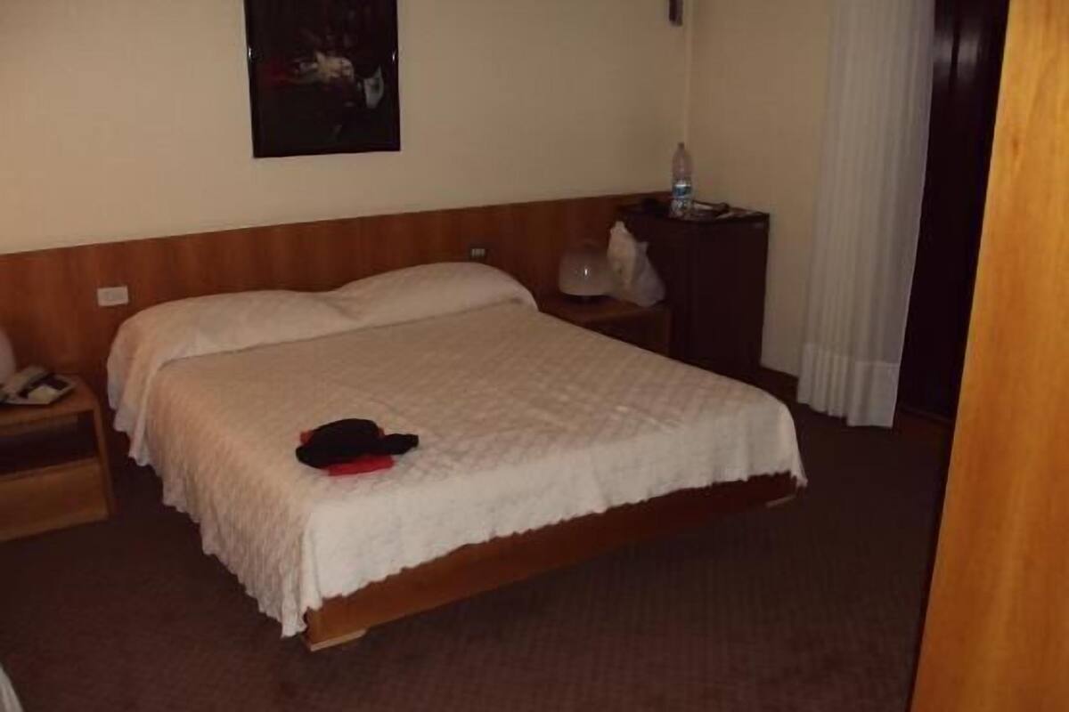 A hotel room with a wooden bed, a nightstand with a lamp, a water bottle, and a painting on the wall.