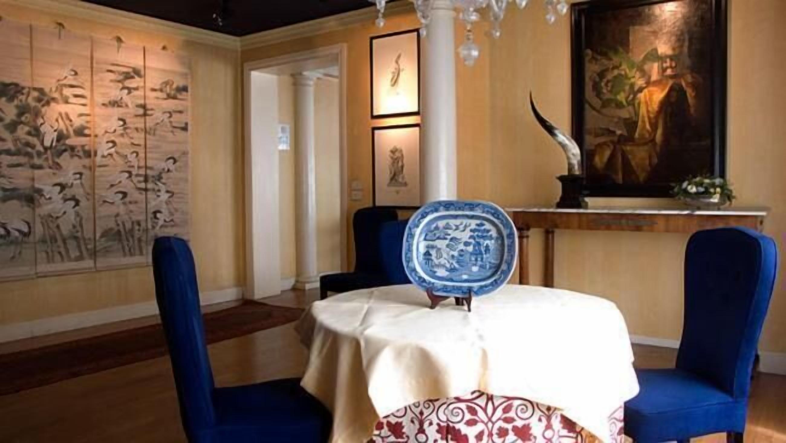 Un comedor con una mesa redonda cubierta por un mantel blanco, dos sillas azules, un plato de cerámica azul y blanco sobre la mesa, y una pared con un cuadro grande.