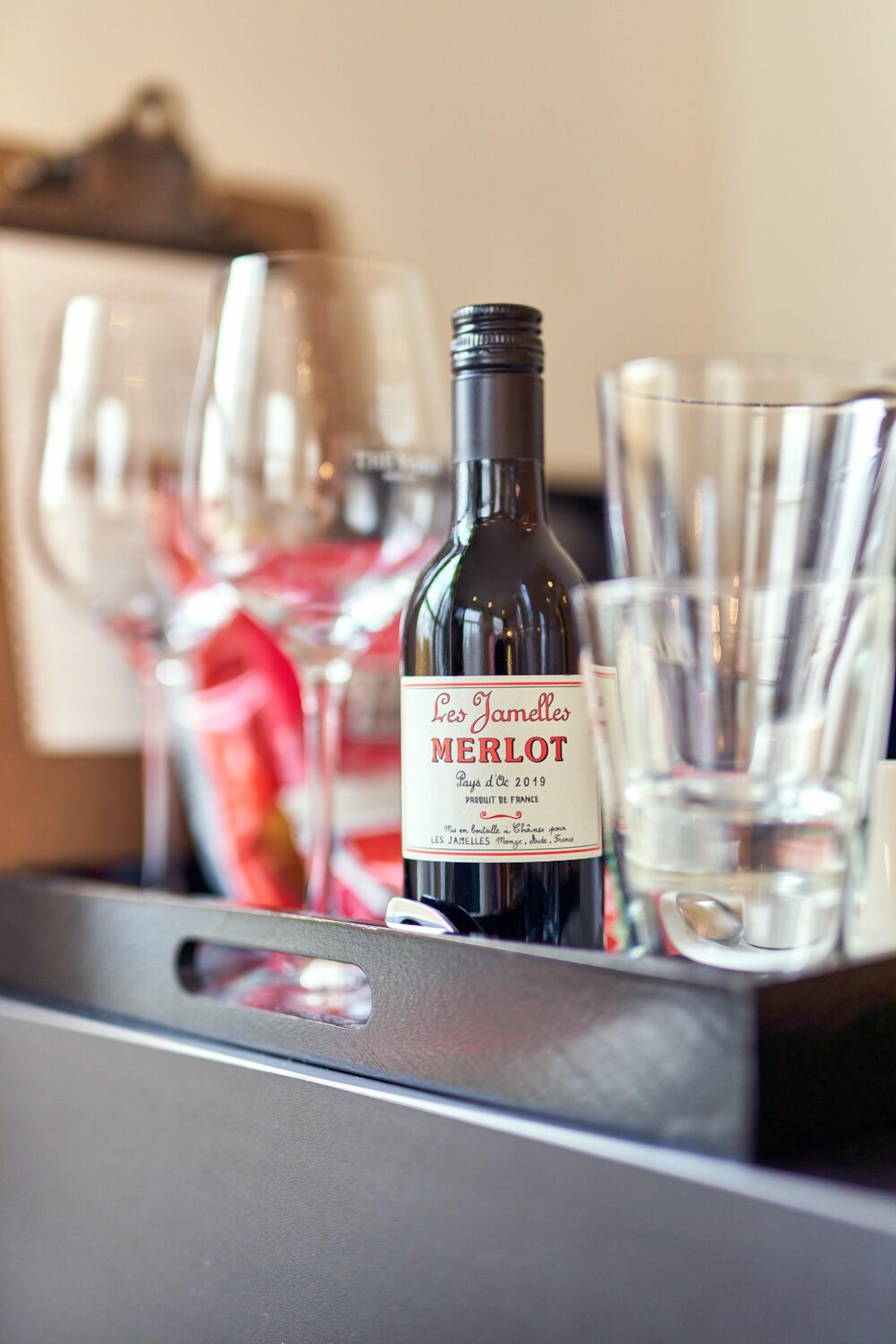 A bottle of Les Jamelles Merlot wine, a glass, and a tray with glasses.