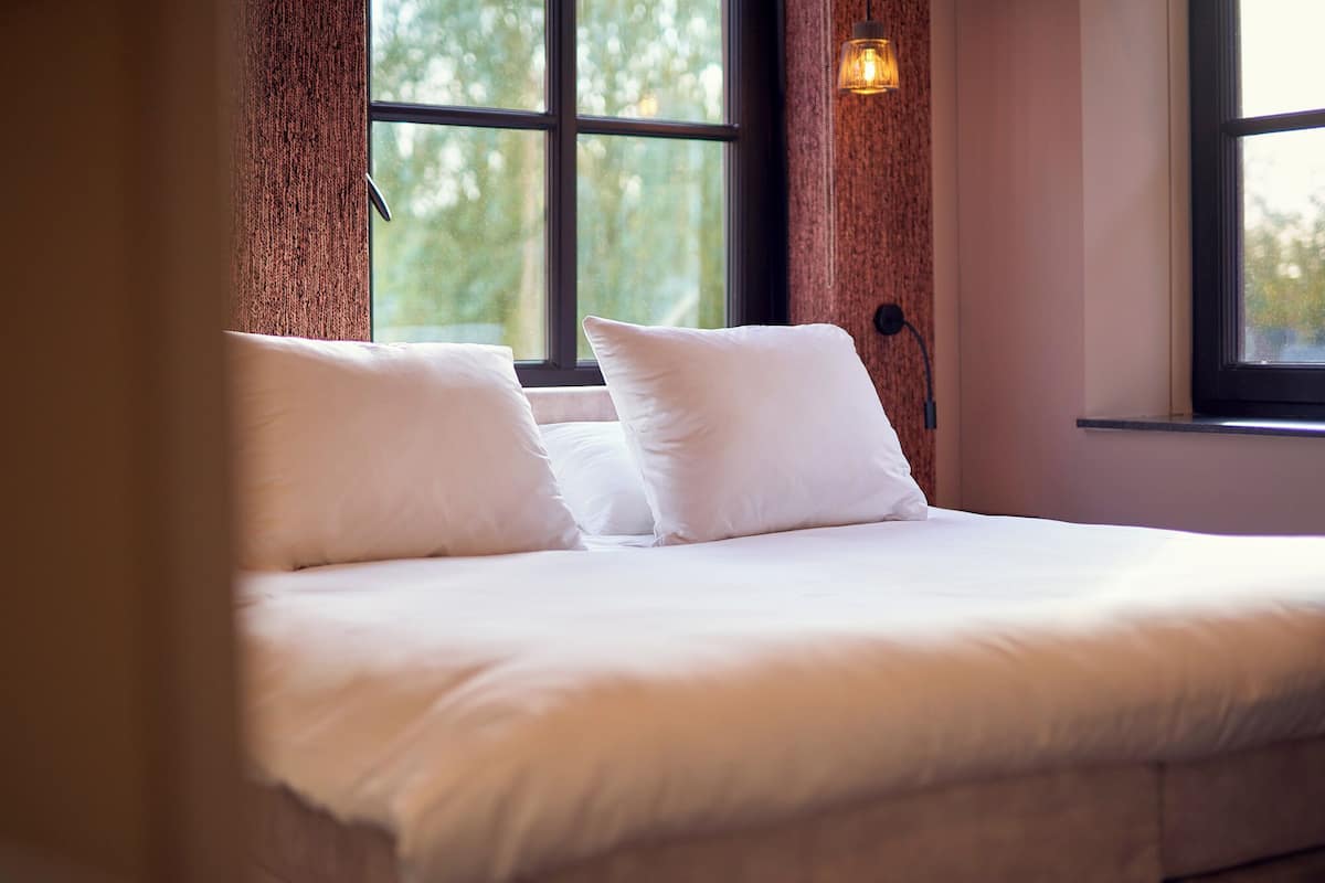 Una cama con sábanas blancas y dos almohadas, una ventana con vistas a árboles y una lámpara fijada a la pared.