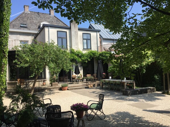 Un patio con mesas al aire libre, una pérgola y un edificio de grandes ventanales y tejado de tejas.