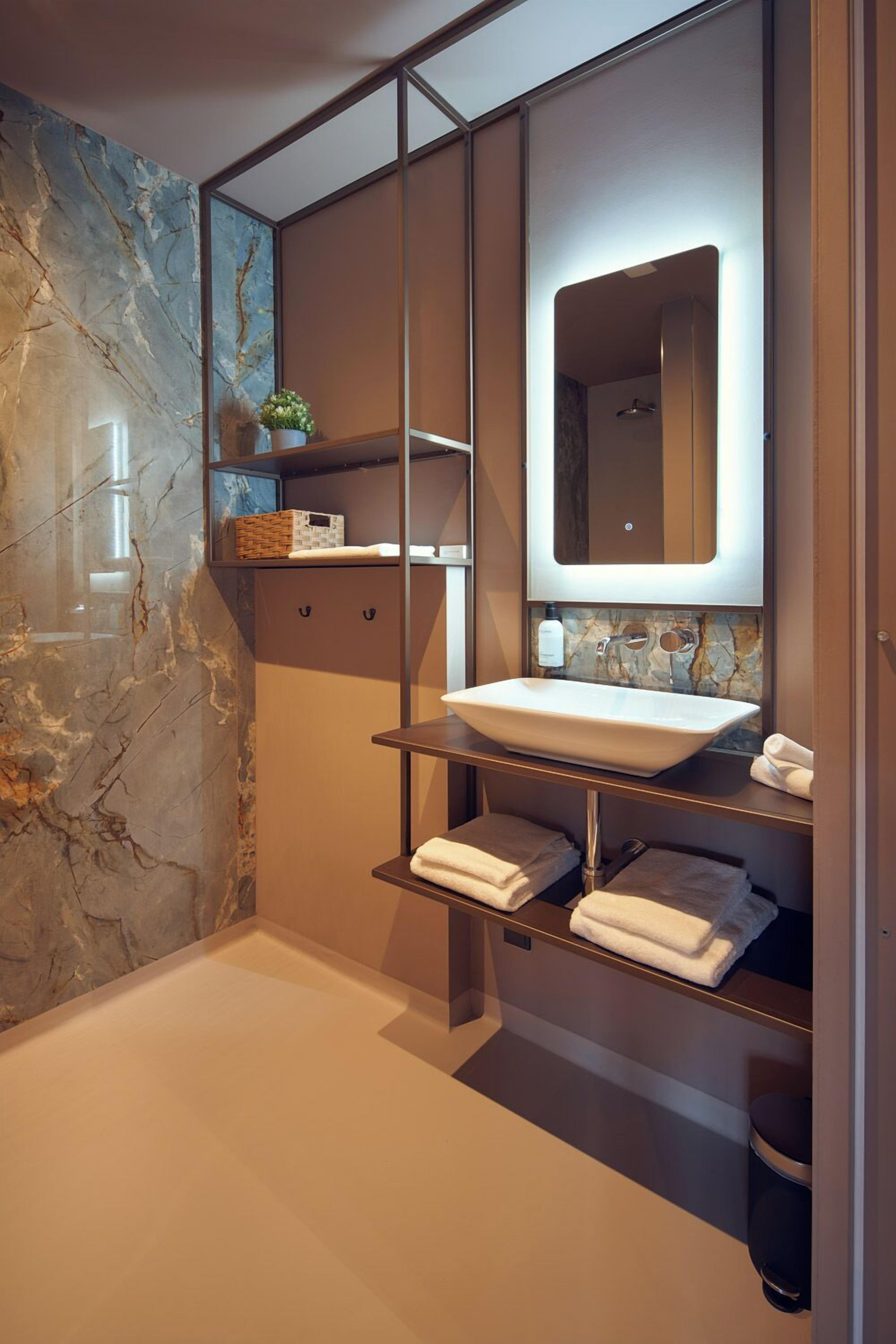 A modern bathroom with a marble wall, a large mirror, a white sink, and shelves with towels.