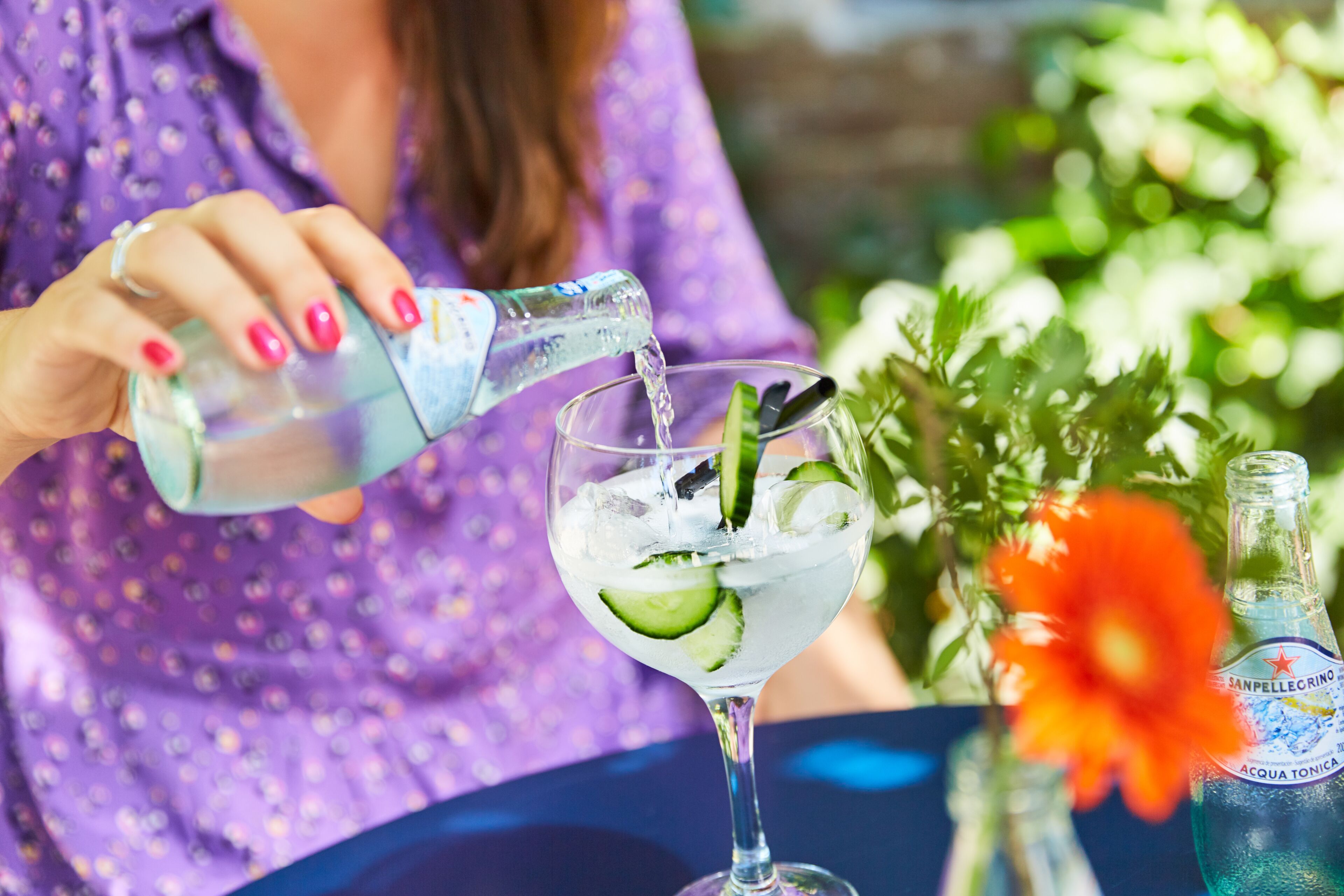 A person pouring a clear liquid into a glass with ice and cucumber slices.