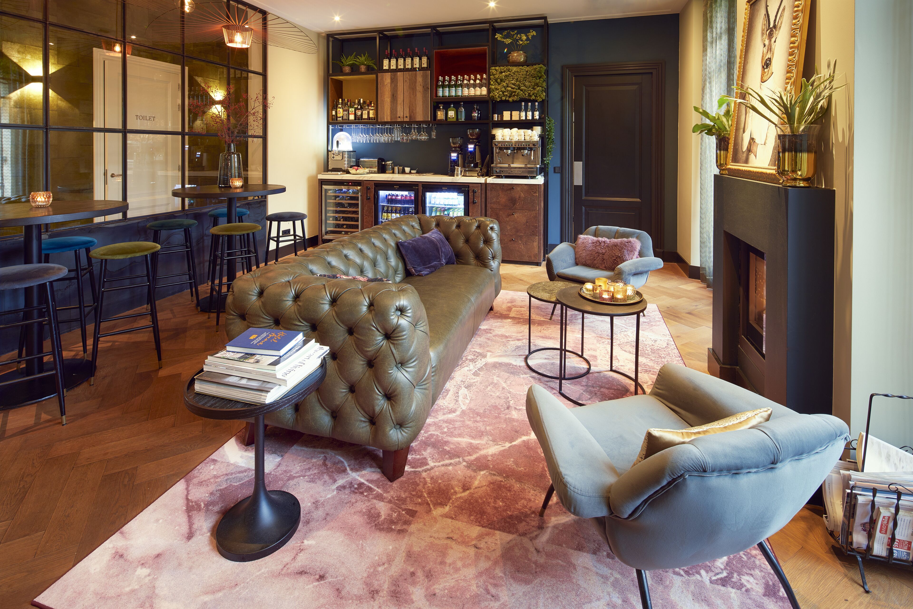 A modern living room with a leather sofa, a blue armchair, and a coffee table. There is a bar area with stools and a minibar.