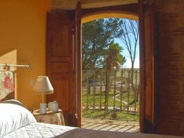 Una habitación con una cama, una mesita de noche con lámpara y vista a un jardín por una puerta abierta.