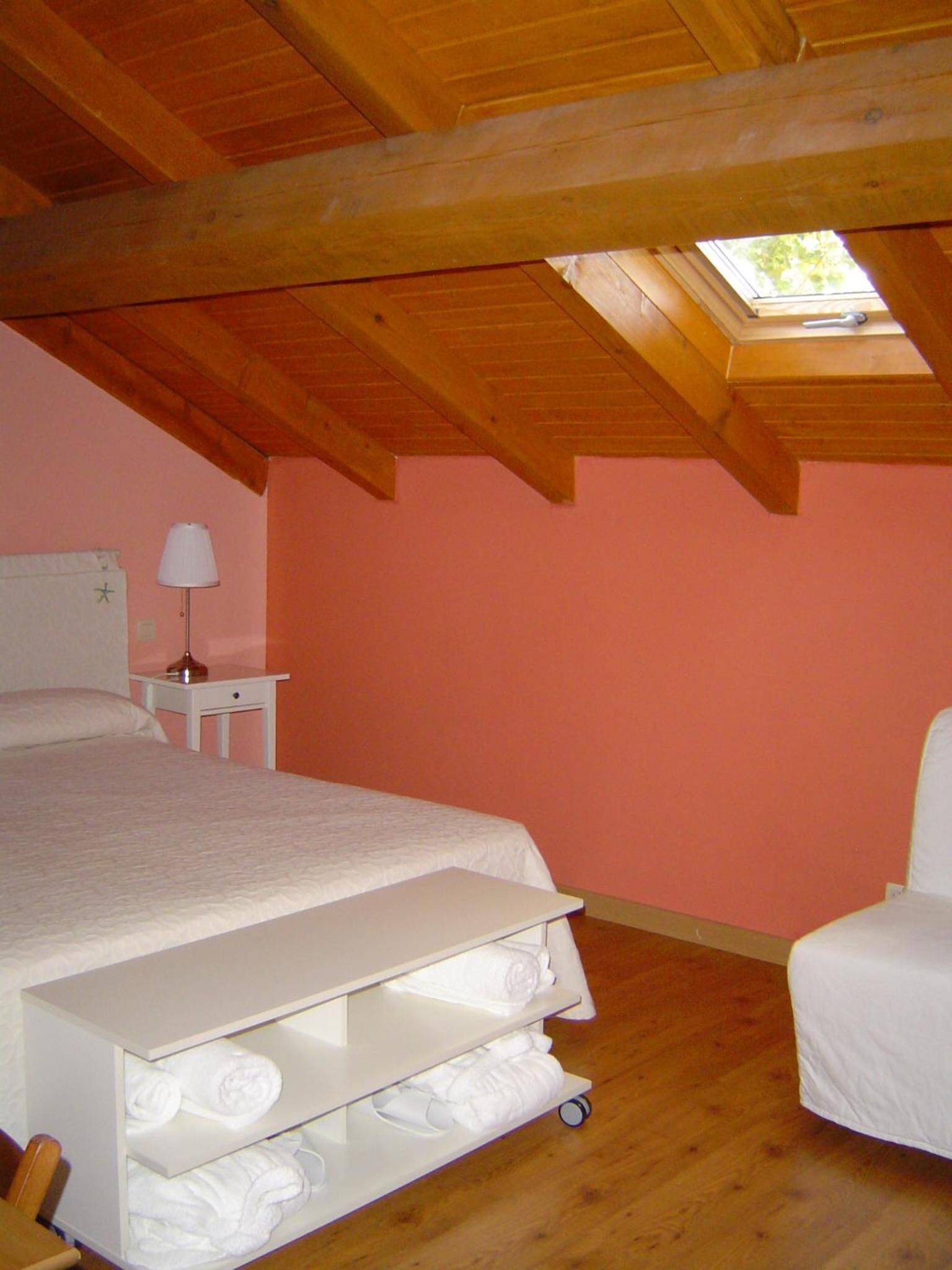 A bedroom with a sloped ceiling, a bed, a nightstand, a lamp, a chair, and a shelf with towels.
