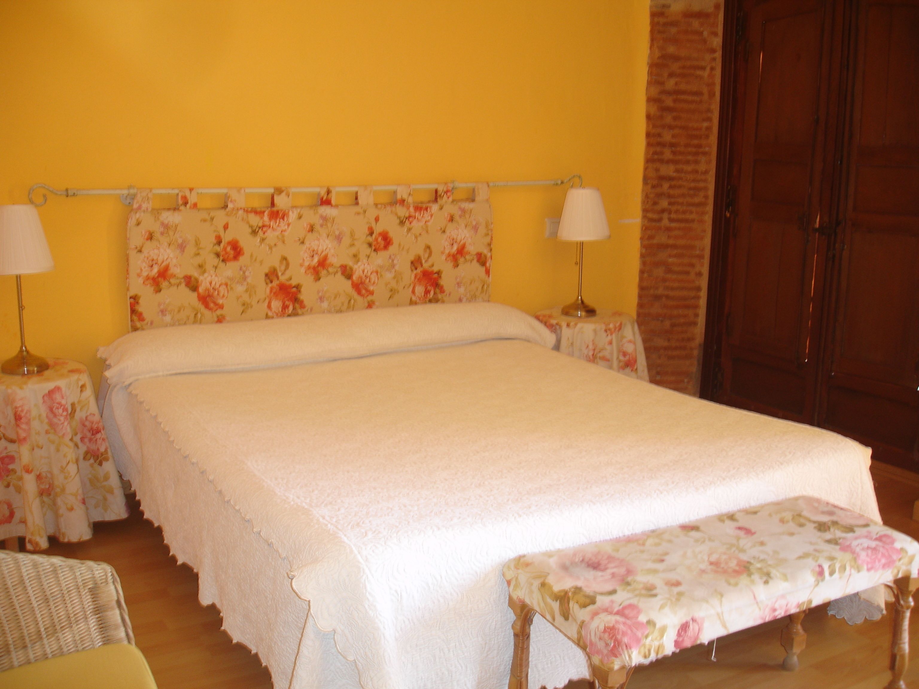 Una cama con cabecera floral, dos mesitas de noche con lámparas, y un banco al pie de la cama.