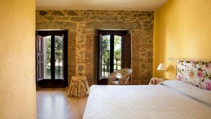 Un dormitorio con pared de piedra, cama con cabecera floral, una silla de mimbre y una pequeña mesa con mantel floreado.