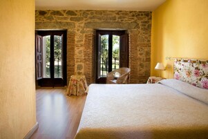 Un dormitorio con pared de piedra, cama con cabecera floral, una silla de mimbre y una pequeña mesa con mantel floreado.