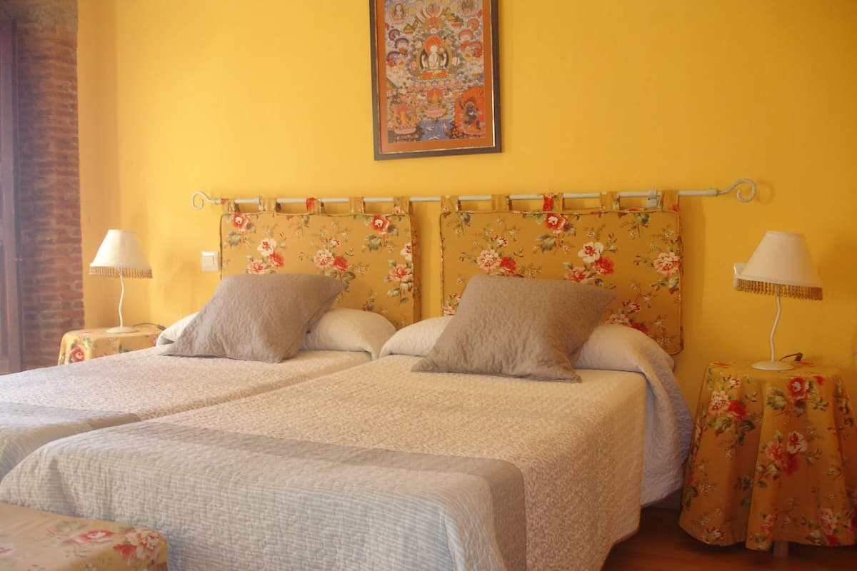 A double bed with floral headboard and two pillows, a nightstand with a lamp, and a small table with a floral tablecloth.
