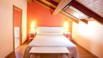 A narrow, single-room hotel bedroom with a sloped ceiling, a bed, two bedside tables, and a small window.