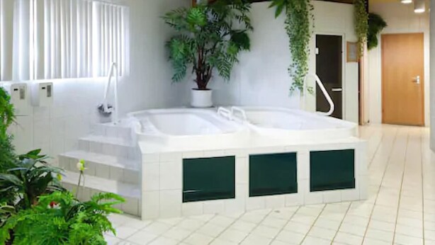A modern bathroom with a freestanding bathtub, white tiles, wooden beams, and potted plants. - Sporthotel Malchow (Malchow)