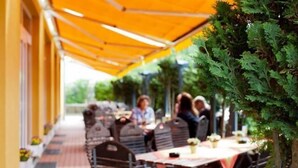 An outdoor dining area with tables and chairs under a yellow canopy, surrounded by greenery. - Sporthotel Malchow (Malchow)