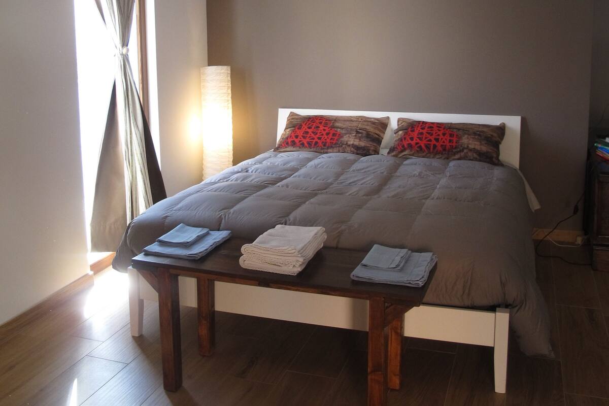 A neatly made bed with a grey duvet, two pillows, and a bedside table with folded towels.